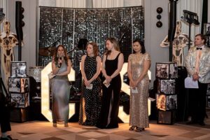 Four women in fancy dresses on stage, one with a microphone