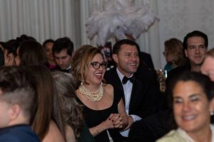 Looking through the crowd at a women in glasses a pearl necklace smiling next to man in tuxedo