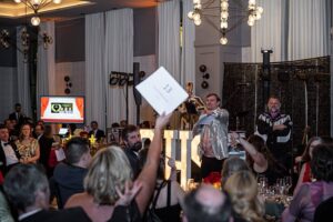 View from the audience of an auction, with the auctioneer on stage in a silver shiny coat